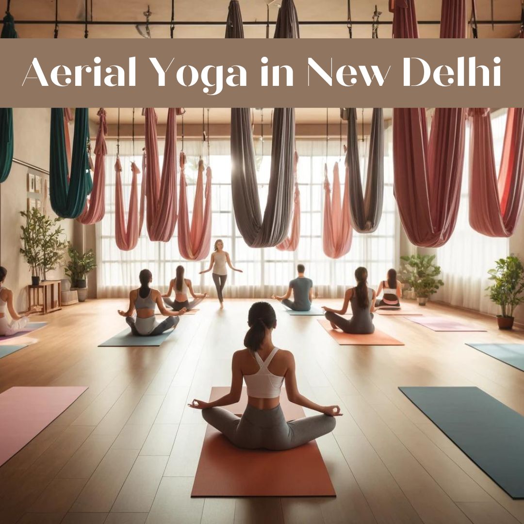 A serene aerial yoga class at The Pink Lotus Academia in New Delhi, featuring participants using colorful hammocks suspended from the ceiling while performing various poses. The studio has soft, natural lighting with large windows, and the instructor is guiding the class in a tranquil, inviting atmosphere with plants and neatly arranged yoga mats in the background.